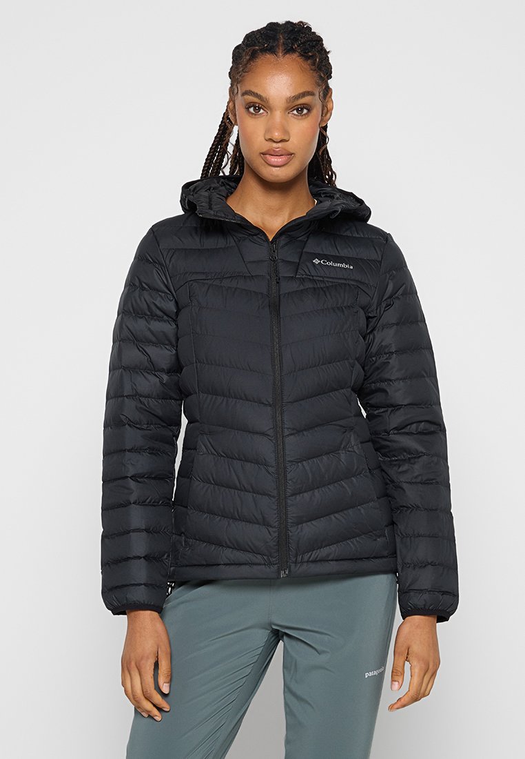 Kobieta z warkoczami, ubrana w czarną kurtkę puchową z kapturem i szare spodnie sportowe, stoi na jednolitym tle.