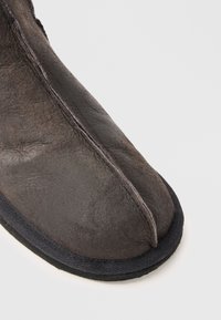 Detailní záběr hnědé kožené tabí boty s designem rozděleného prstu a černým lemem na bílém pozadí.