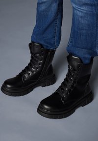 Bottines en cuir noir avec une semelle robuste et texturée, dotées d'œillets métalliques pour le laçage et d'une fermeture éclair latérale pour un accès facile. Portées avec un jean bleu.