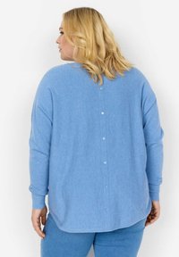 Šviesiai mėlynas ilgomis rankovėmis marškinėliai, pasižymintys švelnia tekstūra ir eilute mygtukų ant nugaros. Suapvalintas apatinis kraštas ir šiek tiek laisvas pritaikymas.