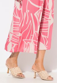 Beige gewebte Mules mit einem niedrigen Absatz und offenem Zeh, kombiniert mit einem rosa Kleid, das ein kühnes weißes abstraktes Muster aufweist.