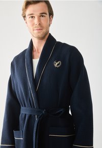 Navy Waffelpique-Bademantel mit kontrastierendem goldenen Piping, mit einem Emblem auf der Brust, zwei vorderen Taschen und einem Bindegürtel.