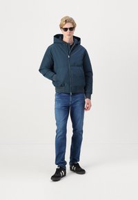 Chaqueta acolchada de color verde oscuro con capucha, cremallera frontal, dos bolsillos inferiores; llevada encima de unos vaqueros azules y zapatillas negras con rayas blancas.