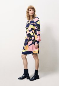 Ärmeldress in Marineblau, Pink, Gelb und grünem abstraktem Muster. Verfügt über ein Wickeldesign, einen Bindegürtel und wird mit schwarzen Ankle Boots kombiniert.