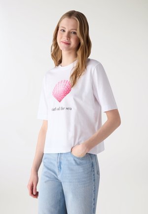 Giovane donna con lunghi capelli biondi che indossa una maglietta bianca con una conchiglia rosa e la scritta "perla del mare", abbinata a jeans azzurri.