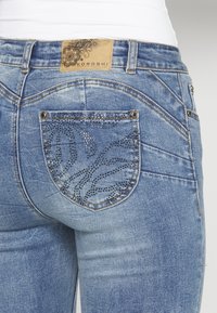 Jeans i denim med ljusblå tvätt, dekorerad bakficka med rhinestone-mönster och en märkesläderetikett vid midjan.