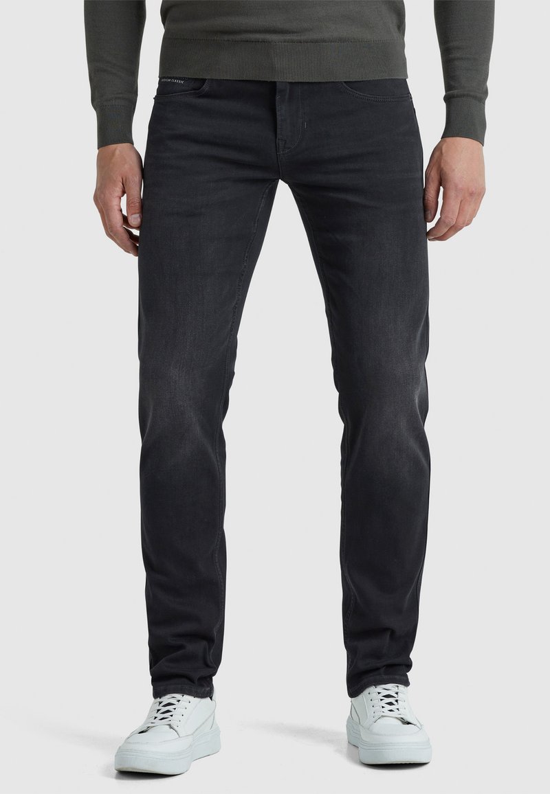 Uomo che indossa jeans neri slim-fit, una maglietta a maniche lunghe grigia e sneakers bianche, in piedi contro uno sfondo chiaro e neutro.