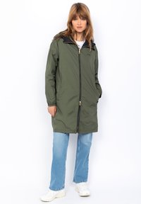 Cappotto lungo imbottito verde oliva con colletto nero, cerniera frontale e due tasche laterali, indossato sopra jeans blue e sneakers bianche.