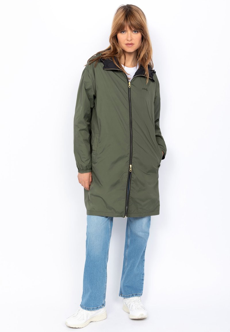 Manteau long en olive avec un col noir, fermeture éclair sur le devant et deux poches latérales, porté sur un jean bleu et des baskets blanches.