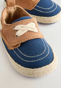 Des espadrilles en toile bleue présentent une sangle en tissu marron avec un détail de nœud blanc, des coutures blanches et une semelle en jute tressé pour une texture.
