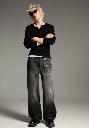 Persona giovane con capelli biondi e occhiali da sole, in piedi con le braccia incrociate, indossa una maglia nera a maniche lunghe, jeans a gamba larga scoloriti e scarpe nere.