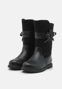 Mustat saappaat, joissa on yhdistelmä kiiltävää nahkaa ja pehmeää mokkaa, polvipituiset, pyöreä nerdo, koristeelliset nauhat ja kukka-akcentit.