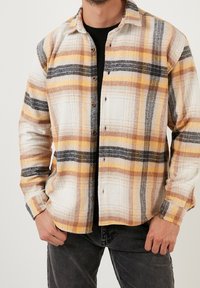 Camisa de cuadros de manga larga en colores blanco, amarillo, marrón y negro. Presenta cierre de botones y un ajuste holgado con una tela texturizada.