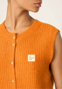 Geverfd oranje gebreide vest met een ronde halslijn, voorzien van een voorste knoopsluiting en een vierkant merkembleem op de borst.