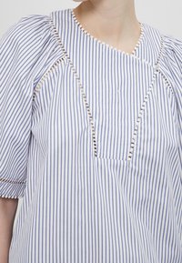 Blouse à rayures en coton blanc et bleu avec un col rond. Dispose de manches bouffantes, d'une bordure décorative et d'un détail de poche avant.