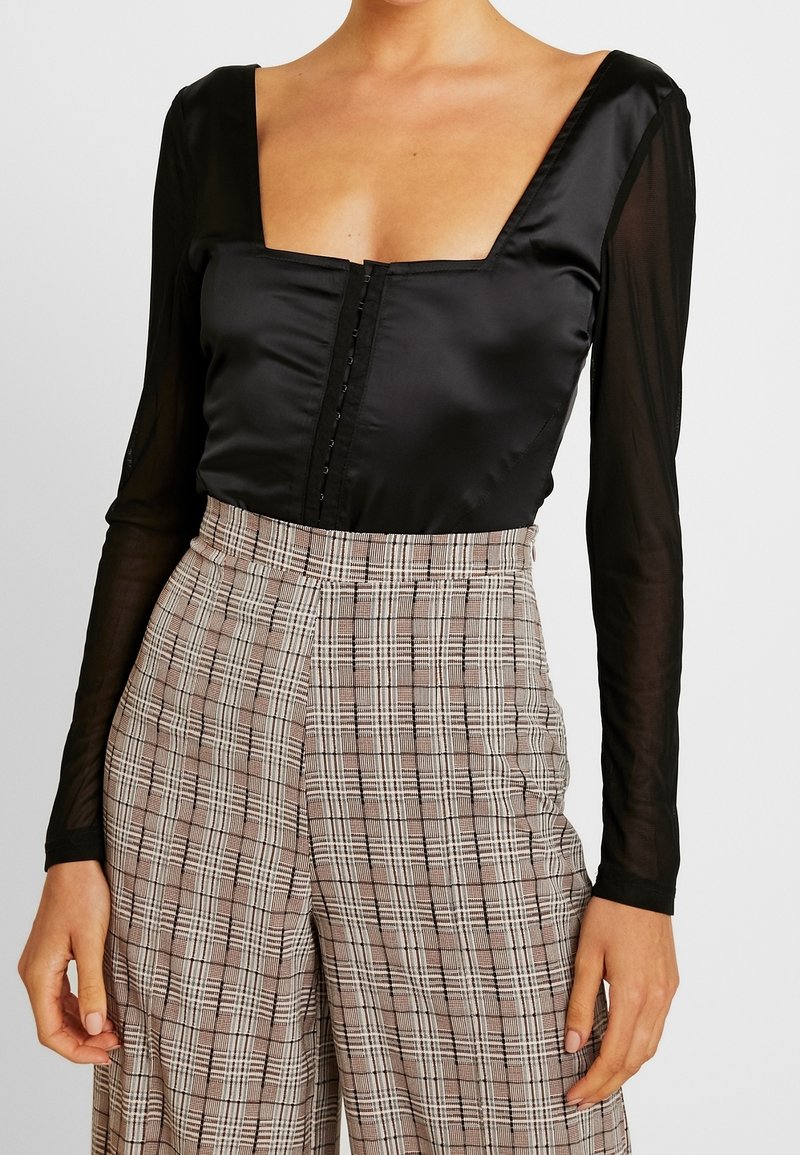 Sort satin langærmet top med firkantet udskæring og knapdetaljer, kombineret med højtaljede ternede bukser i beige og sort mønster.