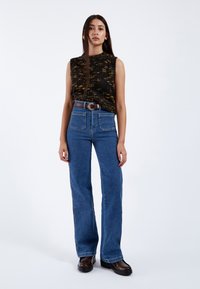 Top de punto sin mangas en verde oscuro y amarillo, combinado con pantalones de mezclilla acampanados de tiro alto en azul y un cinturón marrón. Textura suave y bolsillos visibles.