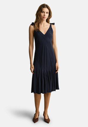 Frau trägt ein ärmelloses, marineblaues Midikleid mit Bindeträgern und V-Ausschnitt, kombiniert mit braunen spitzen High Heels, steht vor weißem Hintergrund.