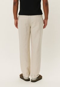 Pantalon en lin beige à coupe décontractée, doté de deux poches arrière et de revers roulés. Porté avec des chaussures de couleur foncée.