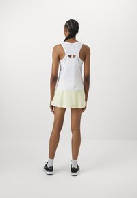 Top blanco sin mangas con detalle recortado en la parte posterior, combinado con una falda amarilla clara. La modelo lleva zapatos negros y calcetines blancos.