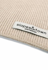 Tessuto a coste beige con etichetta tessuta che mostra "slopes&town AMSTERDAM." Il materiale ha una texture morbida e un motivo consistente.