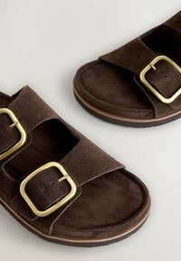 Sandalias de ante marrón con correas anchas y grandes hebillas cuadradas doradas sobre un fondo blanco.