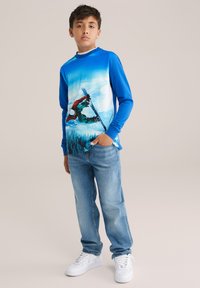 Mėlyna ilgomis rankovėmis palaidinė su slidinėjimo grafika, derinama su šviesiai mėlynomis džinsais ir baltais sportbačiais, turinti laisvą siluetą ir tekstūruotą audinį.