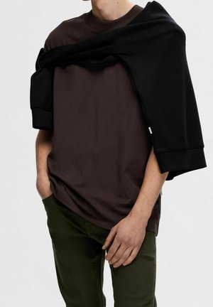 Brązowa koszulka z okrągłym dekoltem, noszona z czarną bluzą narzuconą na ramiona. Oliwkowozielone spodnie dopełniają strój.