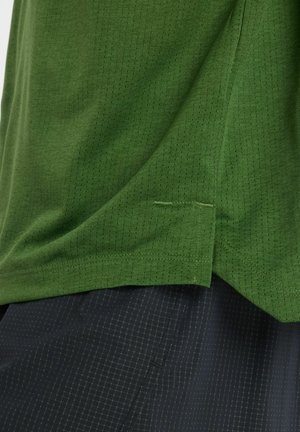 Chemise verte à manches courtes en tissu texturé avec un motif quadrillé subtil, présentant une fente latérale et un ourlet cousu. Associée à un pantalon foncé à motifs.