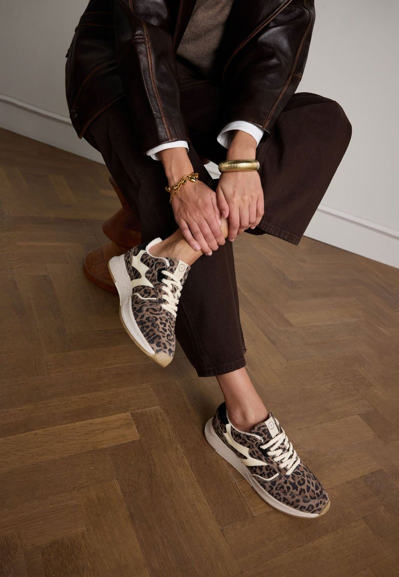 Leopardenmuster-Sneaker mit elfenbeinfarbenen Akzenten, Textil-Obermaterial, sahnefarbenen Schnürsenkeln und Gummisohle. Tragen Sie sie mit brauner Hose auf Holzboden im Fischgrätmuster.