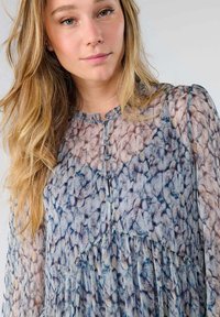 Blusa transparente con escote y botones; estampada en tonos azules y beige; presenta detalles plisados en el dobladillo y las mangas.