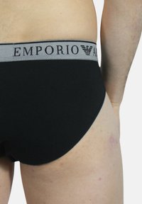 Slip da uomo nero con elastico grigio in vita, caratterizzato dal logo "EMPORIO ARMANI" stampato in nero. Tessuto liscio e design aderente.