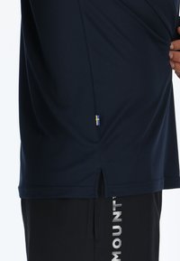 Marineblaues Sportshirt aus strukturiertem Material, mit seitlichem Schlitz und kleinem schwedischen Flaggen-Detail am Saum. Getragen mit schwarzen Shorts, die weißen Text haben.