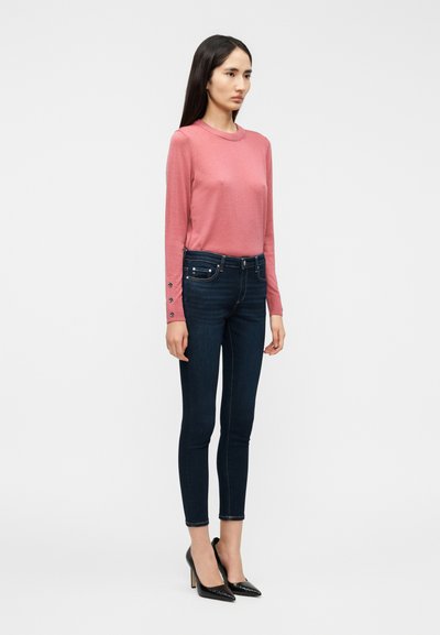 Στενά τζιν σκούρου μπλε χρώματος συνδυασμένα με ροζ ανοιχτό μακρυμάνικη μπλούζα με διακοσμητικά κουμπιά στα μανίκια και μαύρα παπούτσια με ψηλό τακούνι.