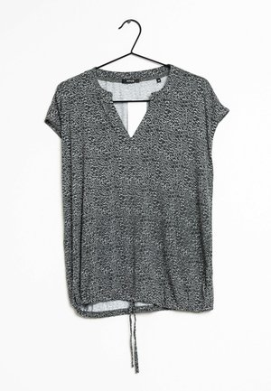 Schwarz-weiß gemusterte Bluse mit kurzen Ärmeln, V-Ausschnitt und verstellbarem Kordelzug am Saum, aufgehängt an einem schwarzen Kleiderbügel vor weißem Hintergrund.