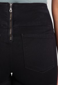 Höga svarta jeans med dragkedja fram, med två bakfickor och subtila sömmar. Slät, stretchig tygstruktur.