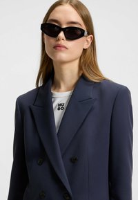 Musta kissakatseinen aurinkolasit kiiltävän kehykset, päällä tummansininen kaksirivinen takki valkoisen paidan kanssa, jossa on teksti "HU GO."