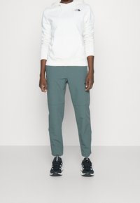 Person som bär en vit The North Face-hoodie, tealbyxor, vita strumpor och svarta Adidas-sneakers stående framför en enfärgad vit bakgrund.