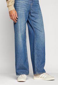 Jean en denim bleu à jambe large avec un lavage délavé, des coutures renforcées et un design classique à cinq poches. Associé à des baskets crème.