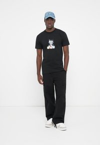 Black cotton t-shirt featuring a cartoon cat graphic, paired with black cargo pants and a light blue cap. Shoes are white with grey accents.