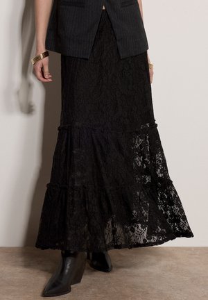 Femme portant une longue jupe en dentelle noire, des bottes noires à bouts pointus, un blazer sombre à fines rayures et des bracelets dorés aux deux poignets.