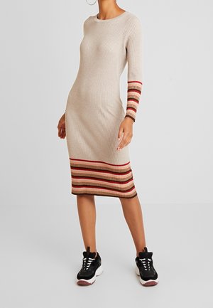 Kobieta w beżowej sukience z prążkowanej dzianiny z wielokolorowymi paskami na mankietach i brzegu, w połączeniu z czarno-białymi masywnymi sneakersami.