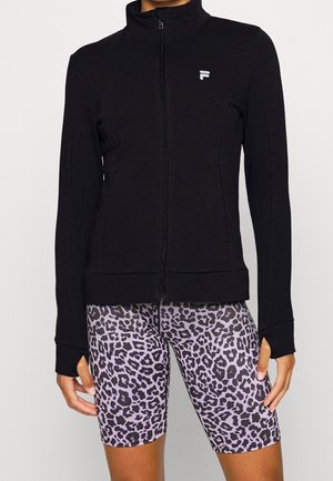 Persoon draagt een zwart jack met rits en duimgaten en paarse, nauwsluitende korte broek met luipaardprint, staand tegen een effen achtergrond.