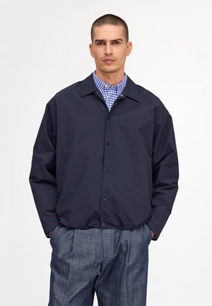 Homme portant une veste bleu marine foncée boutonnée sur une chemise à carreaux bleue et un pantalon gris, debout les mains dans les poches devant un fond blanc.
