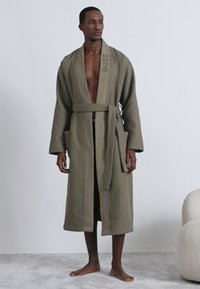 Accappatoio color cachi con trama a nido d'ape, cintura annodata, maniche lunghe e tasche frontali. Presenta un logo nell'area del colletto.