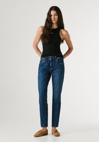 Jauna sieviete ar gariem tumšiem matiem, tērpusies melnā bezpiedurkņu topā, zilās džinsās un brūnās loaferēs, stāvot pretī vienkāršam gaišam fonam.