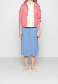 Světle růžová mikina na zip s bílými stahovacími šňůrkami, bílá blůza s vázáním v pase a plisovaná modrá midi sukně s hnědými knoflíky. Béžové tenisky.