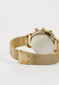 Zegarek z złotą siateczkową bransoletą, wyposażony w stalową kopertę i przezroczysty dekielek. Trzy przyciski oraz regulowane zapięcie dopełniają design.