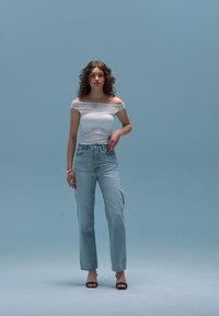 Top bianco scollato, jeans azzurri a vita alta e sandali neri con tacco. La modella è in piedi contro uno sfondo blu uniforme.