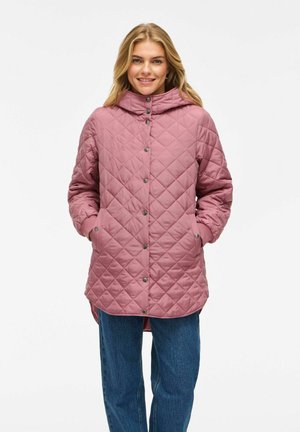 Woman smiling, wearing a dusty pink quilted jacket with snap buttons and hands in pockets, paired with blue jeans against a white background.